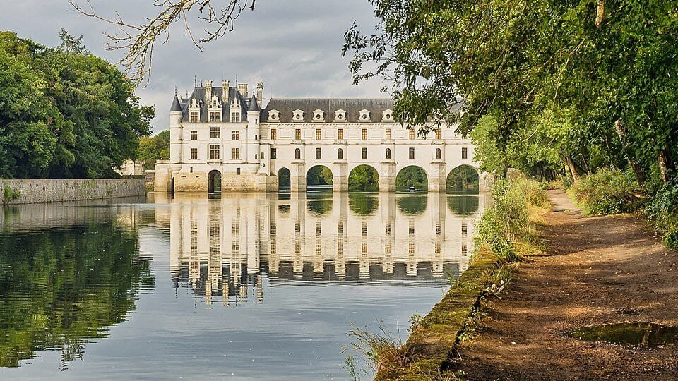 Château Renaissance blanc dont la galerie à arches traverse une rivière, reflété dans l'eau