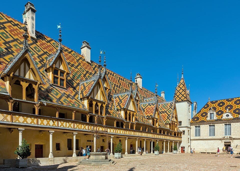 Cour d'un bâtiment médiéval aux toits couverts de tuiles colorées formant des losanges, avec une galerie de bois