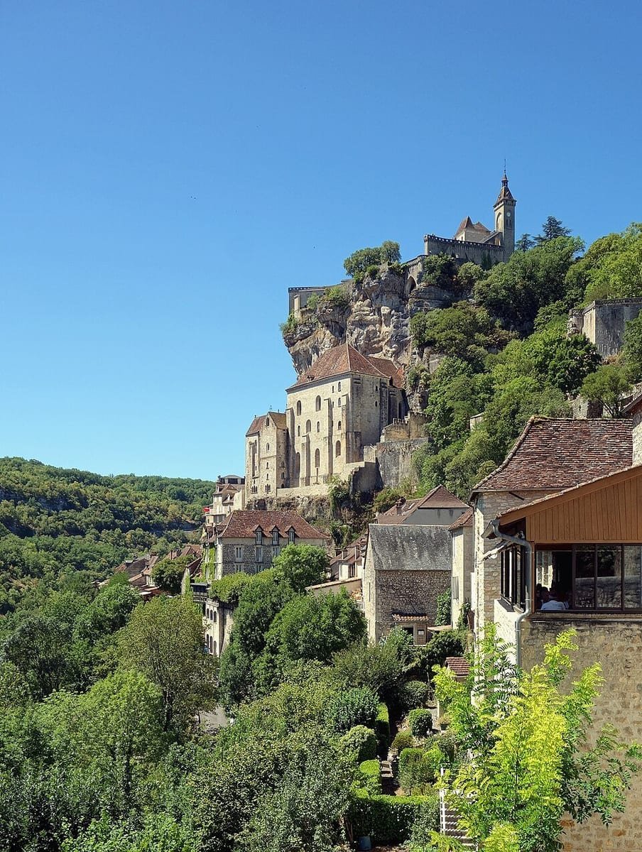 Village de pierre et sanctuaire étagés à flanc de falaise, dominés par un château, au-dessus d'une vallée boisée