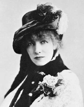 Photographie d'une femme aux cheveux bouclés, coiffée d'un grand chapeau à plumes, une fleur au corsage