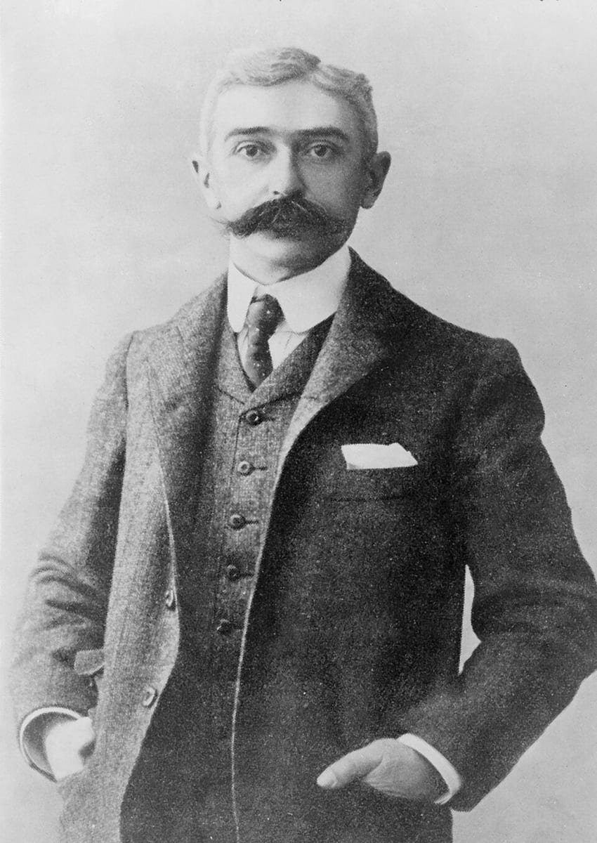 Photographie en noir et blanc d'un homme debout aux cheveux blancs et à grosse moustache, en costume trois pièces, les mains dans les poches