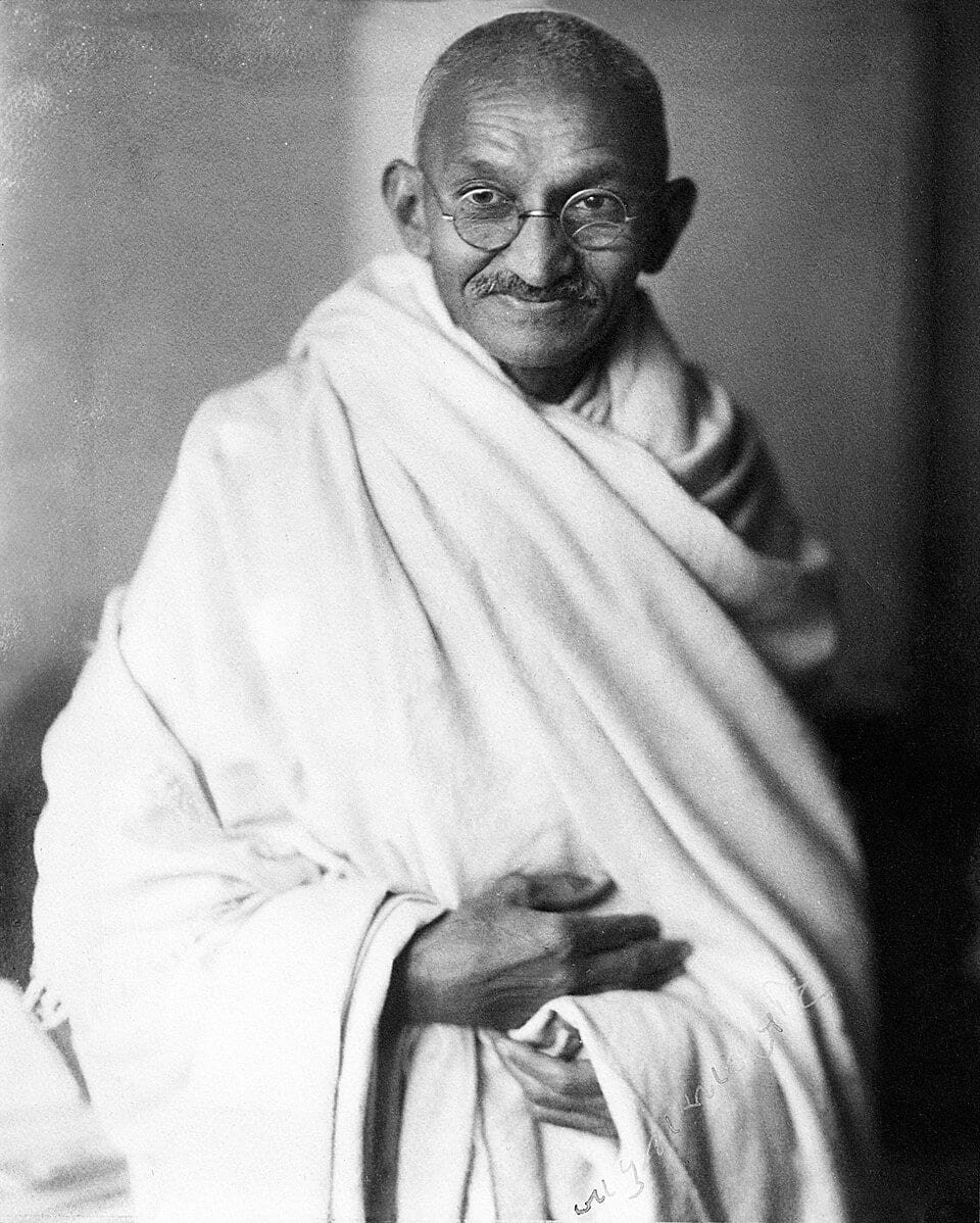 Photographie d'un homme âgé souriant, au crâne rasé et aux lunettes rondes, drapé dans un châle blanc