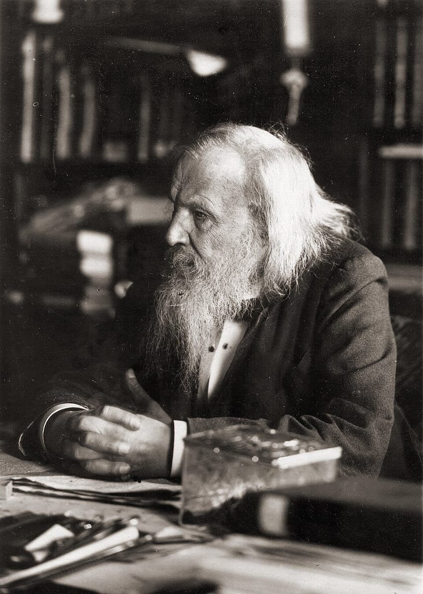 Photographie d'un vieil homme de profil aux longs cheveux et à longue barbe blancs, assis à un bureau encombré de papiers